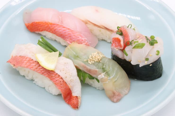 皿に盛られた5貫の寿司。手前左に皮付きの魚の握り、手前中央に緑がかった魚の握り、手前右に軍艦巻き、奥に2貫の握り寿司が並べられています。それぞれの寿司には、薬味やレモンが添えられています。