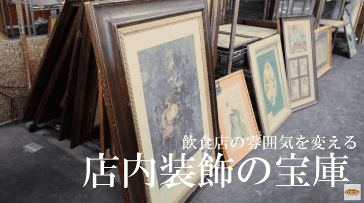飲食店向けの絵画や額縁が多数並べられた倉庫のような空間。手前には大きな額縁に入った絵画が立てかけられており、奥には様々なサイズの絵画や額縁が積まれている。「飲食店の雰囲気を変える 店内装飾の宝庫」という文字が重ねられている。