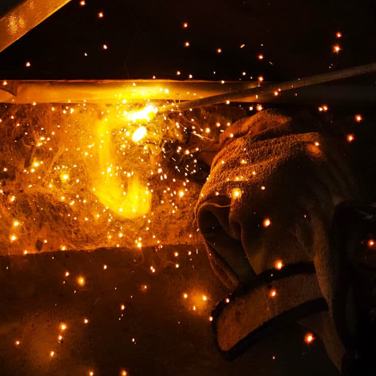 暗い場所で溶接作業が行われており、明るい光と火花が飛び散る様子と作業者の手が写っています。