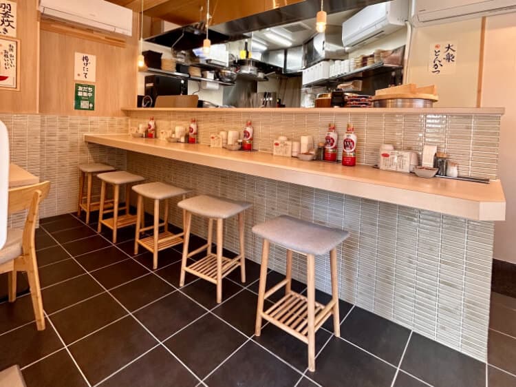 ラーメン屋のカウンター席。木製のカウンターと、タイル張りの壁、そして木製フレームの背もたれのないスツールが並んでいる。