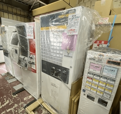 倉庫のような場所で、梱包された中古の券売機が複数台並んでいる写真です。