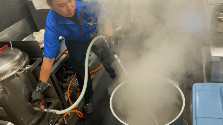 湯気が立ち込めるラーメン店の厨房で、白い調理服を着た職人がガスコンロの上の寸胴鍋を木べらでかき混ぜている様子。手前の作業台にはタイマーや書類、右側の台には焦げ付いた鍋とダシを取り終えた多くの骨が置かれている。